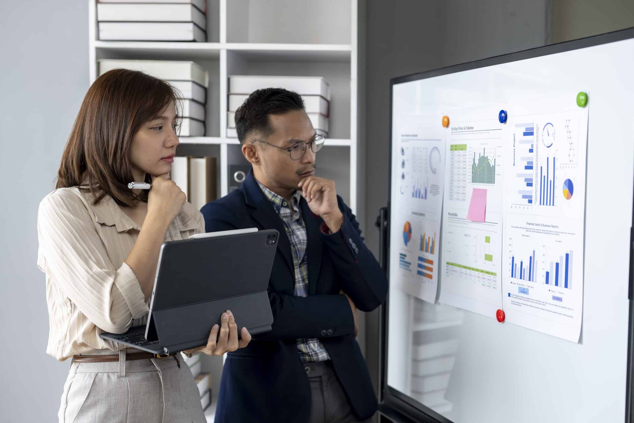 Two people are looking at a white board with graphs and charts. One of them is holding a laptop