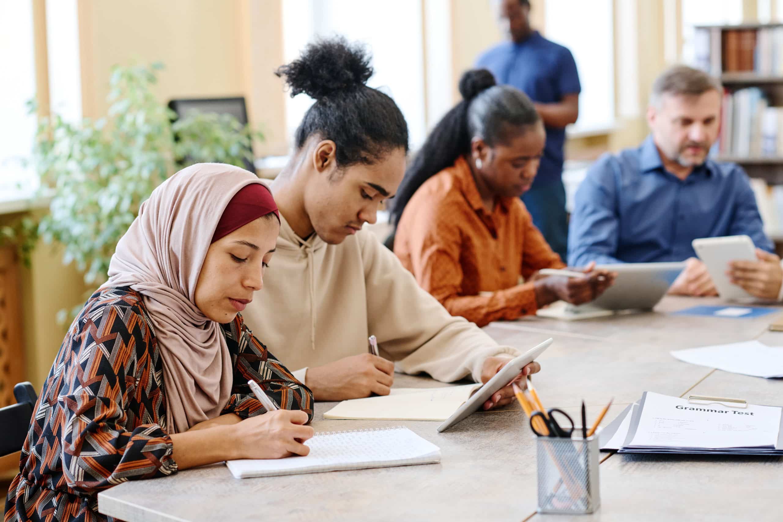 Group of ethnically diverse people having language lesson for immigrants sitting at table doing task with use of digital tablets